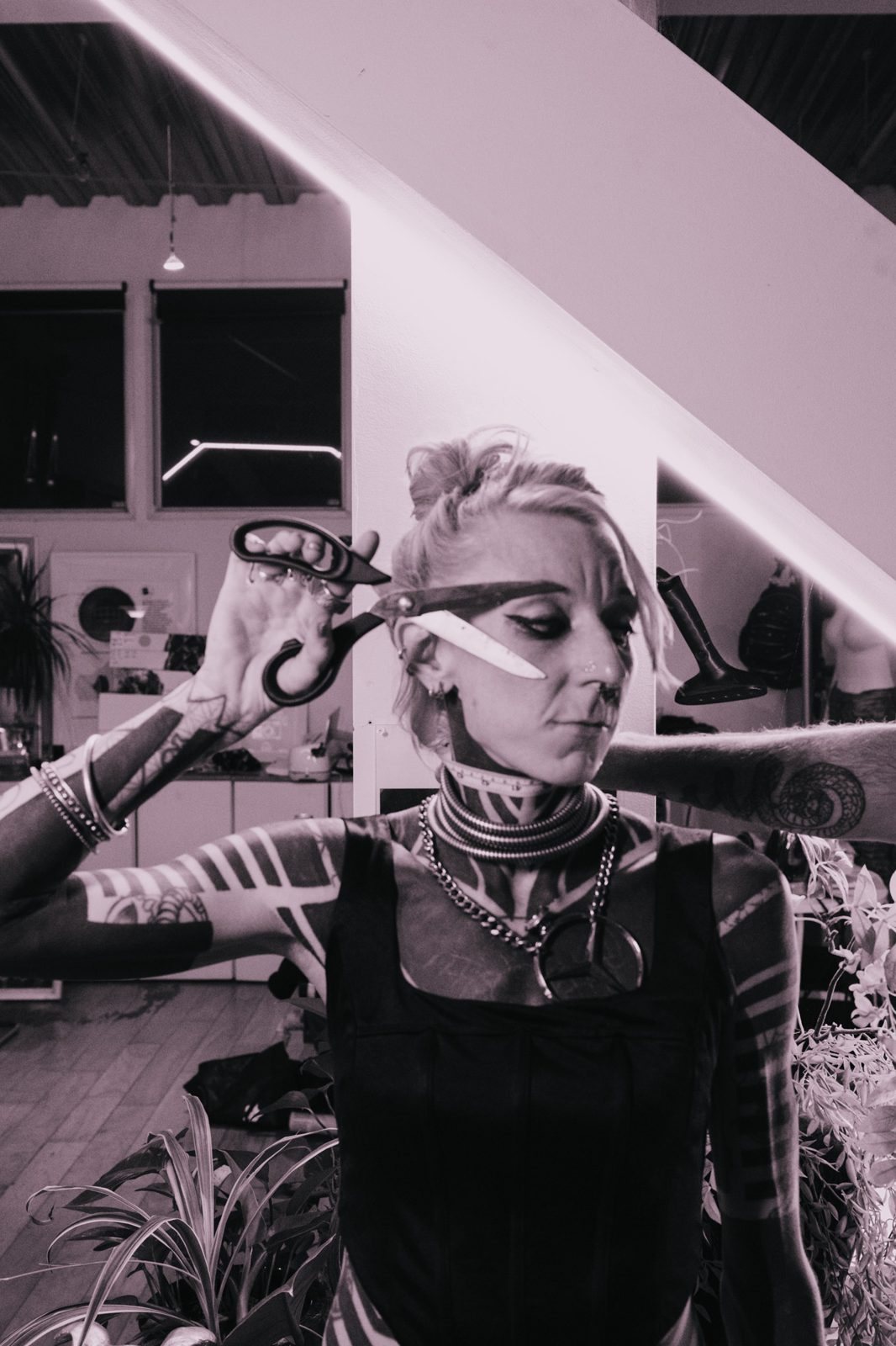 Tattooed designer holding open shears across her eye under a lit staircase