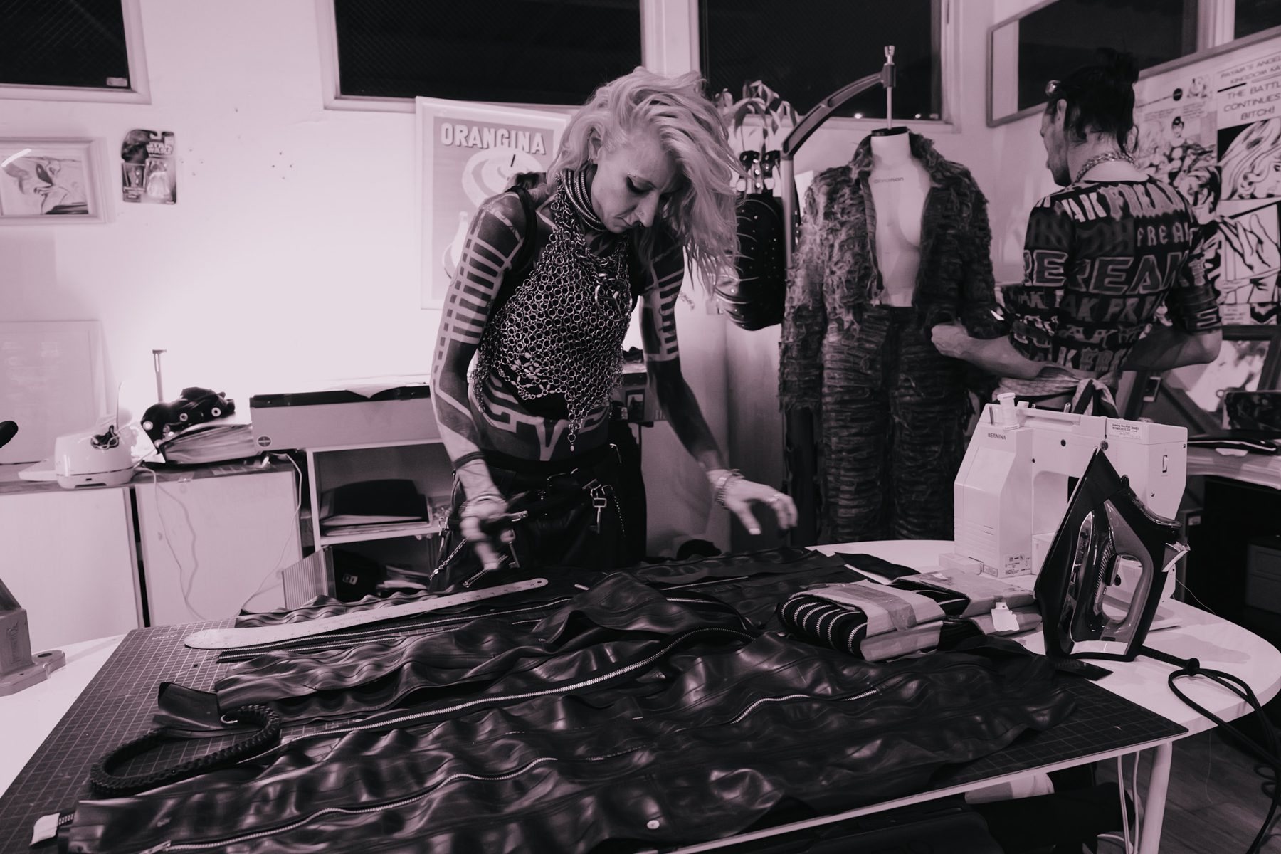 Designer in chainmail laying out zippered leather panels on a cutting table, a distressed suit on a mannequin behind